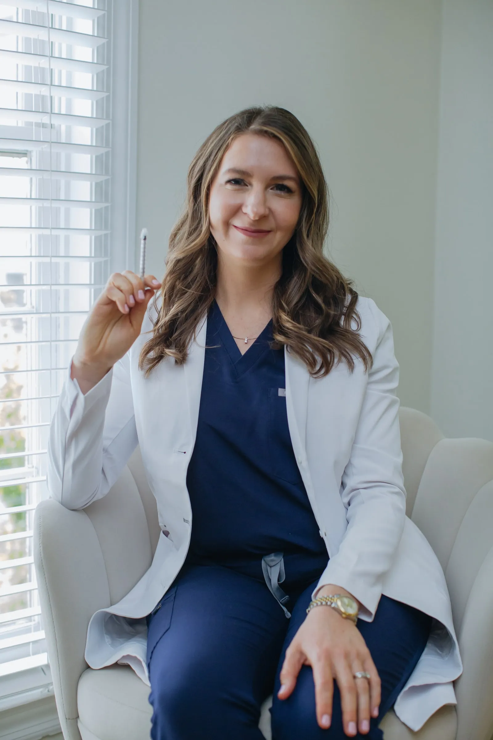 Danielle Wise, NP, in a white coat seated by a window, holding a microneedling pen
